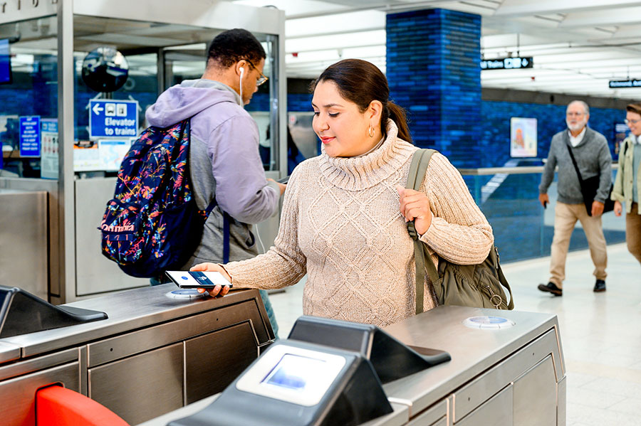 An adult approaches a BART faregate holding their Android phone over the card reader target, using Clipper on their phone to pay for transit.