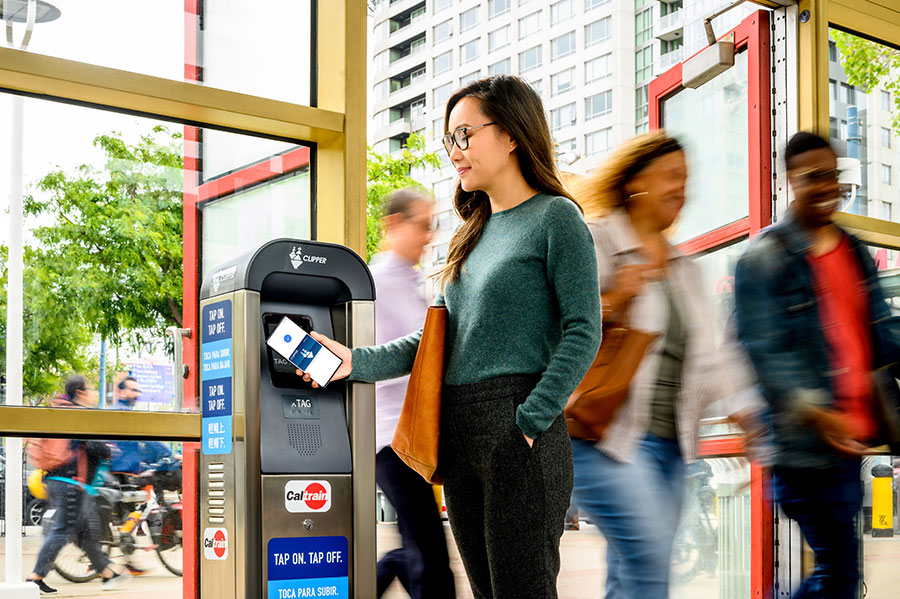 An adult stands in front of a standing Clipper card reader, holding their Android phone up to it, using Clipper on their phone to pay for transit; a “Done” screen is displayed on the phone.